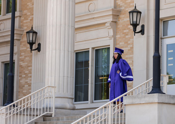 A student in cap and gown standing on the exterior Library steps.